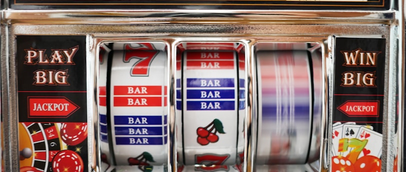 A close-up view of a slot machine with vibrant colors and various gambling symbols such as dice, coins, and playing cards. The reels display a combination of symbols, including bars, cherries, and the number seven. The chrome exterior and colorful details convey the theme of casino and gambling.