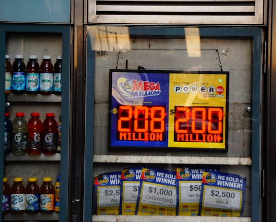 An advertisement display in a store window features lottery jackpot amounts for Mega Millions and Powerball, both showing jackpots of 208 million and 200 million respectively. Below the display, several lottery tickets and promotional posters are visible. To the left of the display, there is a refrigerator stocked with various drinks including different flavors of sports drinks and bottled water.