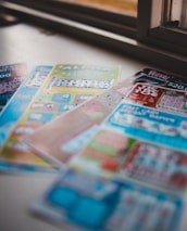 A selection of colorful lottery scratch-off tickets placed on a flat surface near a window. The image has a shallow depth of field, focusing on the tickets while blurring the background. The tickets display various games and prize amounts.