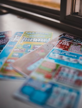 A selection of colorful lottery scratch-off tickets placed on a flat surface near a window. The image has a shallow depth of field, focusing on the tickets while blurring the background. The tickets display various games and prize amounts.