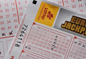 A close-up image of lottery tickets featuring crossed-out numbers, brand logos, and various numbers in a grid format. The tickets have sections for different games, and numbers are marked with black crosses.