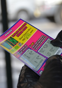 A person wearing black gloves is holding a colorful lottery scratch-off ticket. The ticket includes information about a $1,000,000 guaranteed minimum payout and features multiple games with various prize amounts.