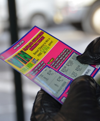 A person wearing black gloves is holding a colorful lottery scratch-off ticket. The ticket includes information about a $1,000,000 guaranteed minimum payout and features multiple games with various prize amounts.