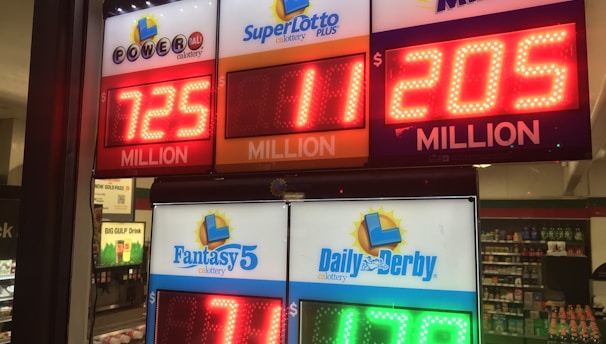 A signboard inside a convenience store displays the current jackpot amounts for various lottery games. There are bright, illuminated numbers in red and green showing the prize amounts: Powerball is at 725 million, Super Lotto Plus at 11 million, Mega Millions at 205 million, Fantasy 5 at 71 thousand, and Daily Derby at 179. In the background, shelves with various drinks and products are visible, along with promotional posters.